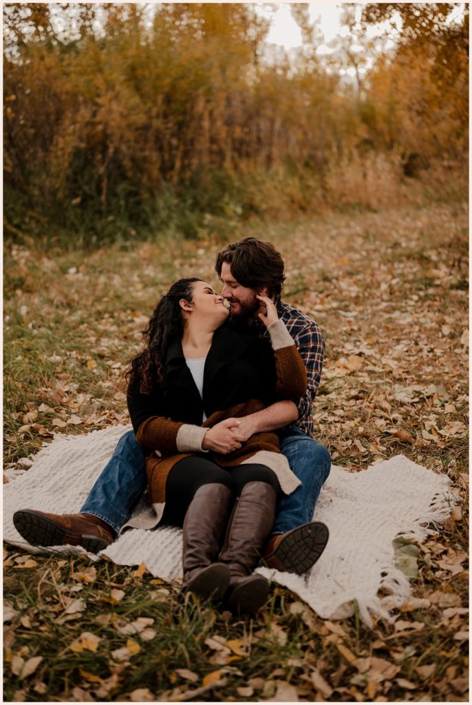 Couple sits on a blanket surrounded by leaves during fall engagement photos in Colorado